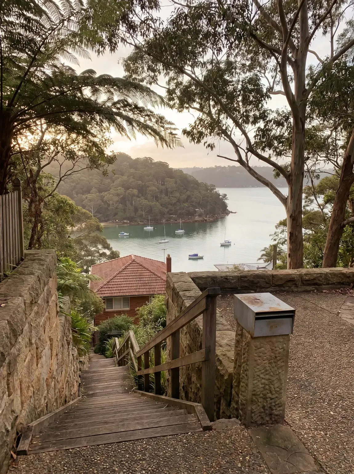 A long private stair flight descending below street level between established gardens toward a house roof and still bay water in Beauty Point