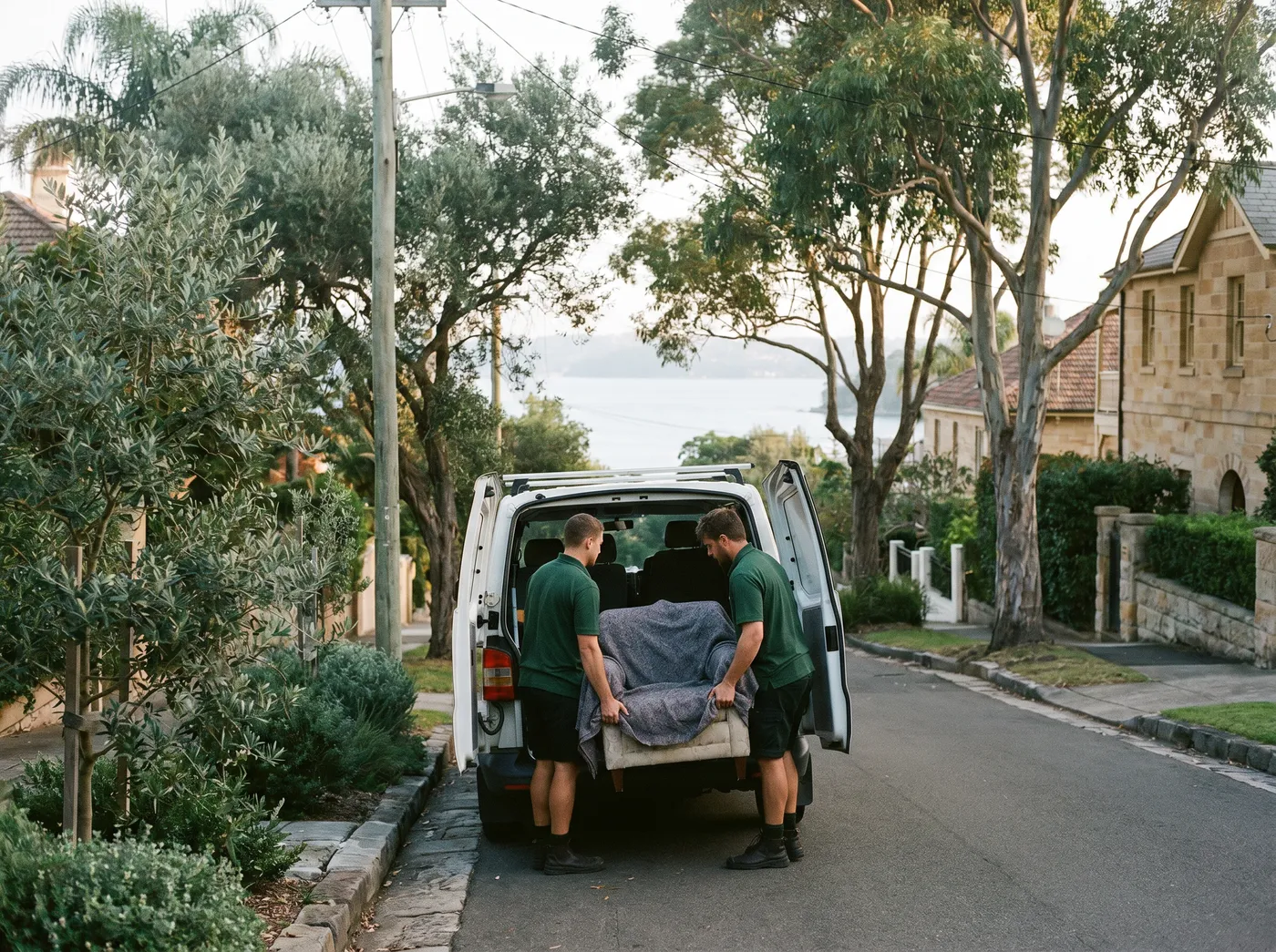 Two crew loading a padded armchair into a plain white van parked on a quiet leafy street above the harbour