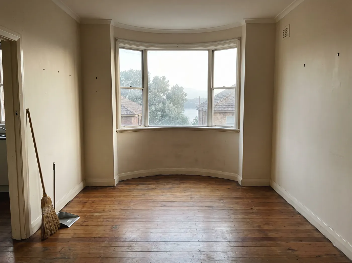 An emptied, freshly swept walk-up apartment room with a curved bay window, bare floorboards and a broom resting by the doorway